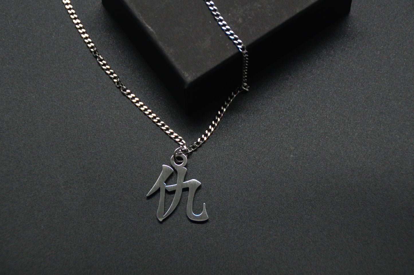 Das Japanische Schriftzeichen für Rache als Edelstahlkette auf einem Schwarzen Hintergrund. Die Kette liegt leicht an einer Kante an.