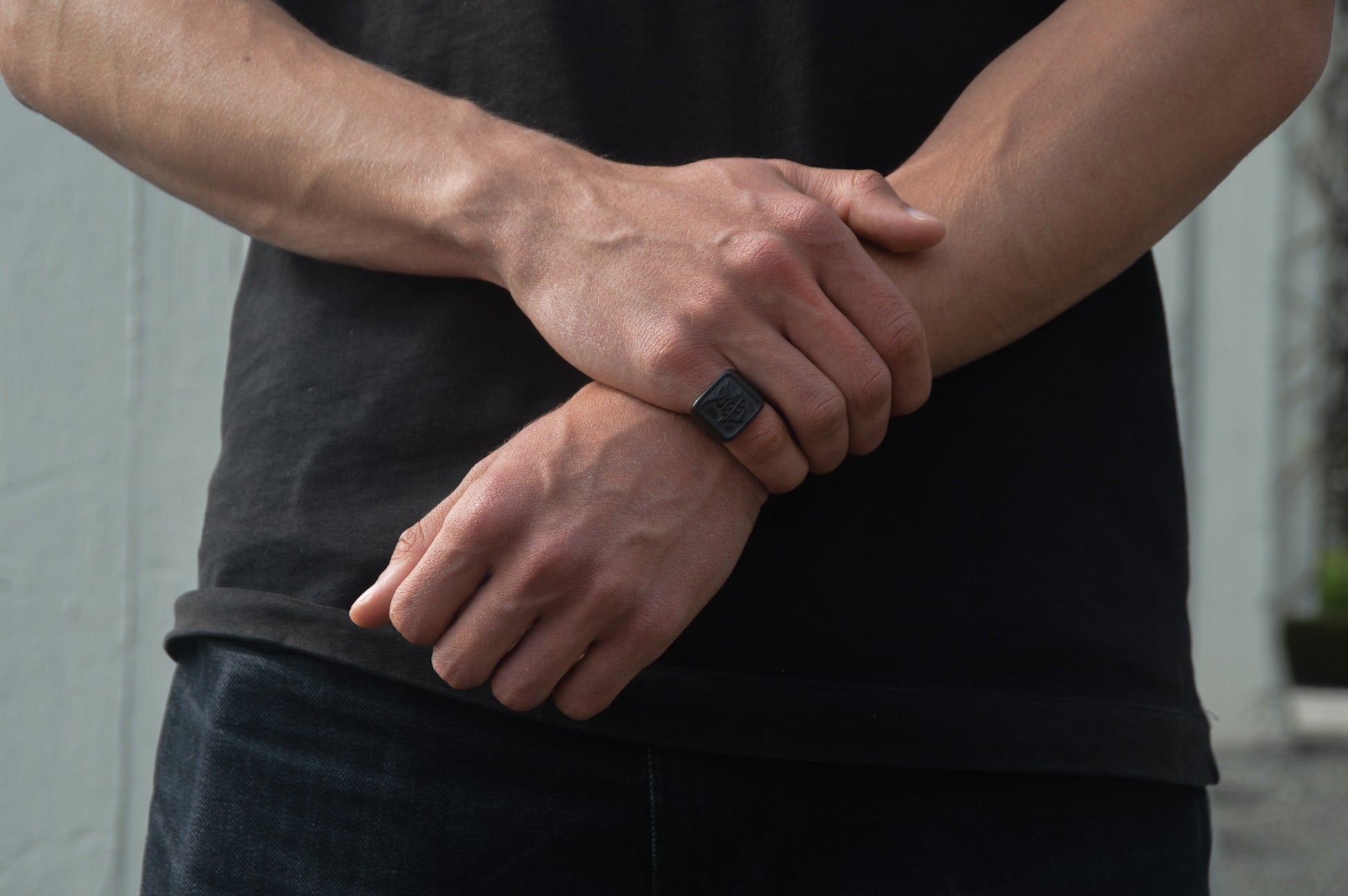 Das Japanische Schriftzeichen für Liebe auf einem Ring. Der Ring ist optisch ein Stempel Ring / Stamp Ring, wobei das Kanji 愛 drauf ist. Zu sehen ist der Schwarze Ring getragen an der rechten Hand auf dem kleinen Finger während der Rechte Arm den linken Arm greift. Frontales Foto