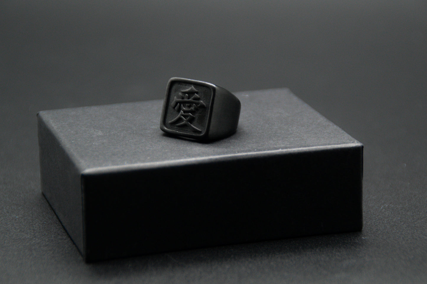 Das Japanische Schriftzeichen für Liebe auf einem Ring. Der Ring ist optisch ein Stempel Ring / Stamp Ring, wobei das Kanji 愛 drauf ist. Zu sehen ist der Schwarze Ring auf einem kleinen Podest von unserer KanjiKraft Box.