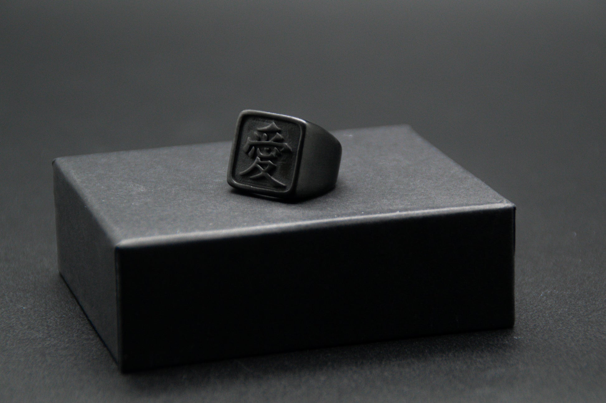 Das Japanische Schriftzeichen für Liebe auf einem Ring. Der Ring ist optisch ein Stempel Ring / Stamp Ring, wobei das Kanji 愛 drauf ist. Zu sehen ist der Schwarze Ring auf einem kleinen Podest von unserer KanjiKraft Box.