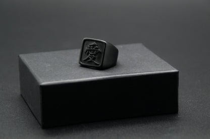 Das Japanische Schriftzeichen für Liebe auf einem Ring. Der Ring ist optisch ein Stempel Ring / Stamp Ring, wobei das Kanji 愛 drauf ist. Zu sehen ist der Schwarze Ring auf einem kleinen Podest von unserer KanjiKraft Box.