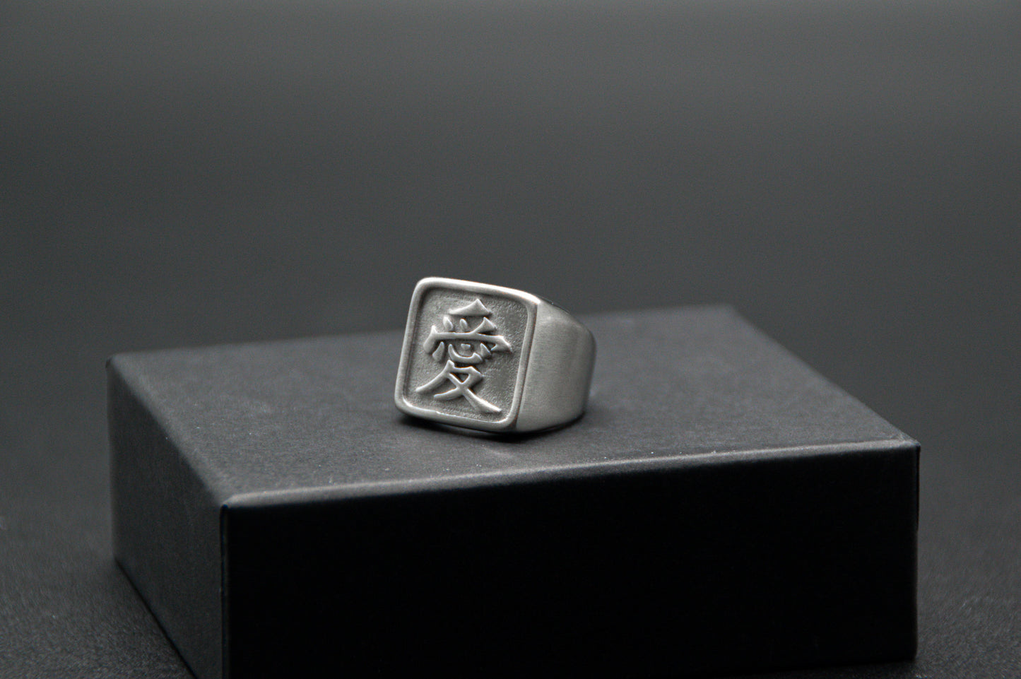 Das Japanische Schriftzeichen für Liebe auf einem Ring. Der Ring ist optisch ein Stempel Ring / Stamp Ring, wobei das Kanji 愛 drauf ist. Zu sehen ist der weiße Ring auf einem kleinen Podest von unserer KanjiKraft Box.