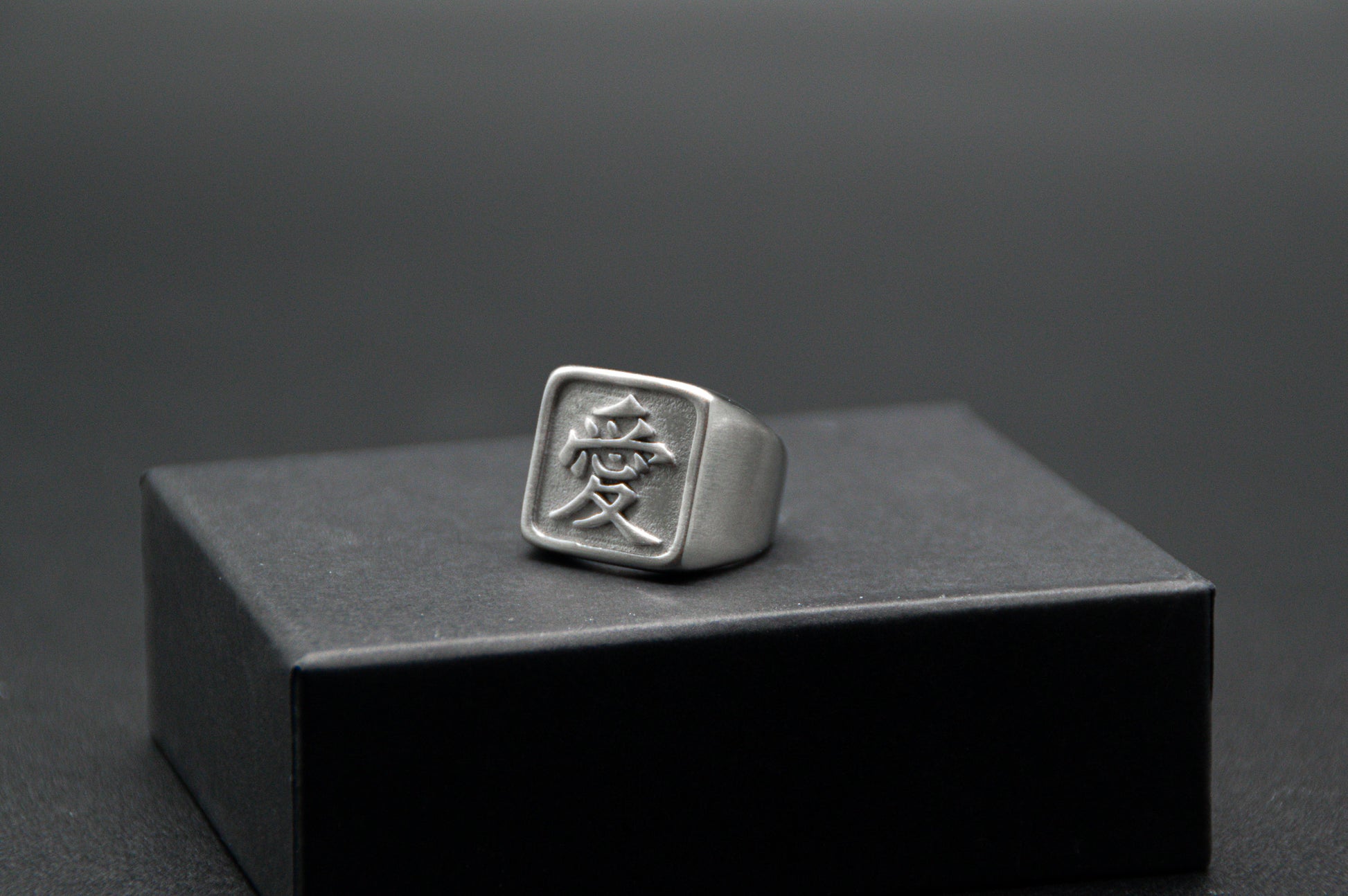 Das Japanische Schriftzeichen für Liebe auf einem Ring. Der Ring ist optisch ein Stempel Ring / Stamp Ring, wobei das Kanji 愛 drauf ist. Zu sehen ist der weiße Ring auf einem kleinen Podest von unserer KanjiKraft Box.