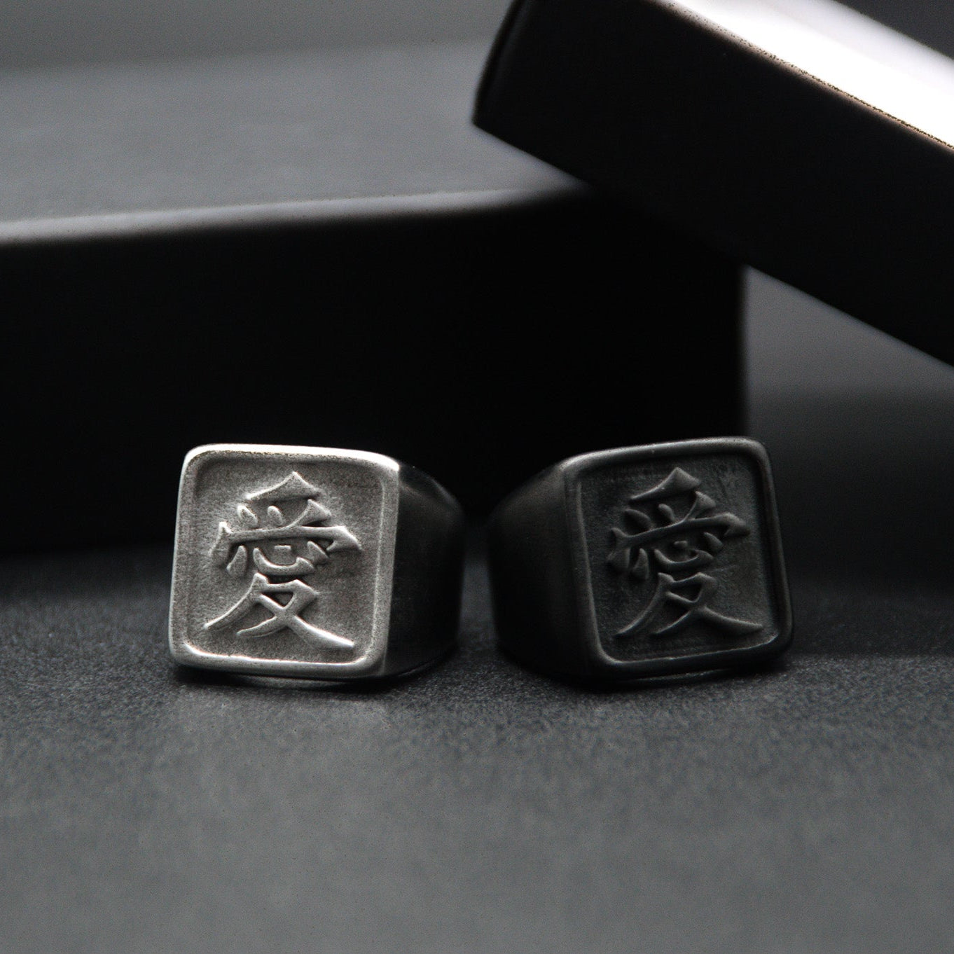 Das Japanische Schriftzeichen für Liebe auf einem Ring. Der Ring ist optisch ein Stempel Ring / Stamp Ring, wobei das Kanji 愛 drauf ist. Beide Ringe sind aus Edelstahl und der linke Ring ist in silberner Farbe und der rechte ist Schwarz und hat eine schwarze Beschichtung.
