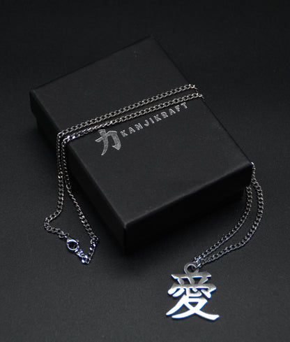 Das Japanische Schriftzeichen (Kanji für Liebe). Edelstahl Halskette mit dem Schriftzeichen 愛 als Anhänger auf einem Schwarzen Hintergrund. Die Kette liegt über der KanjiKraft Geschenkbox. Der Anhänger befindet sich dadrunter.