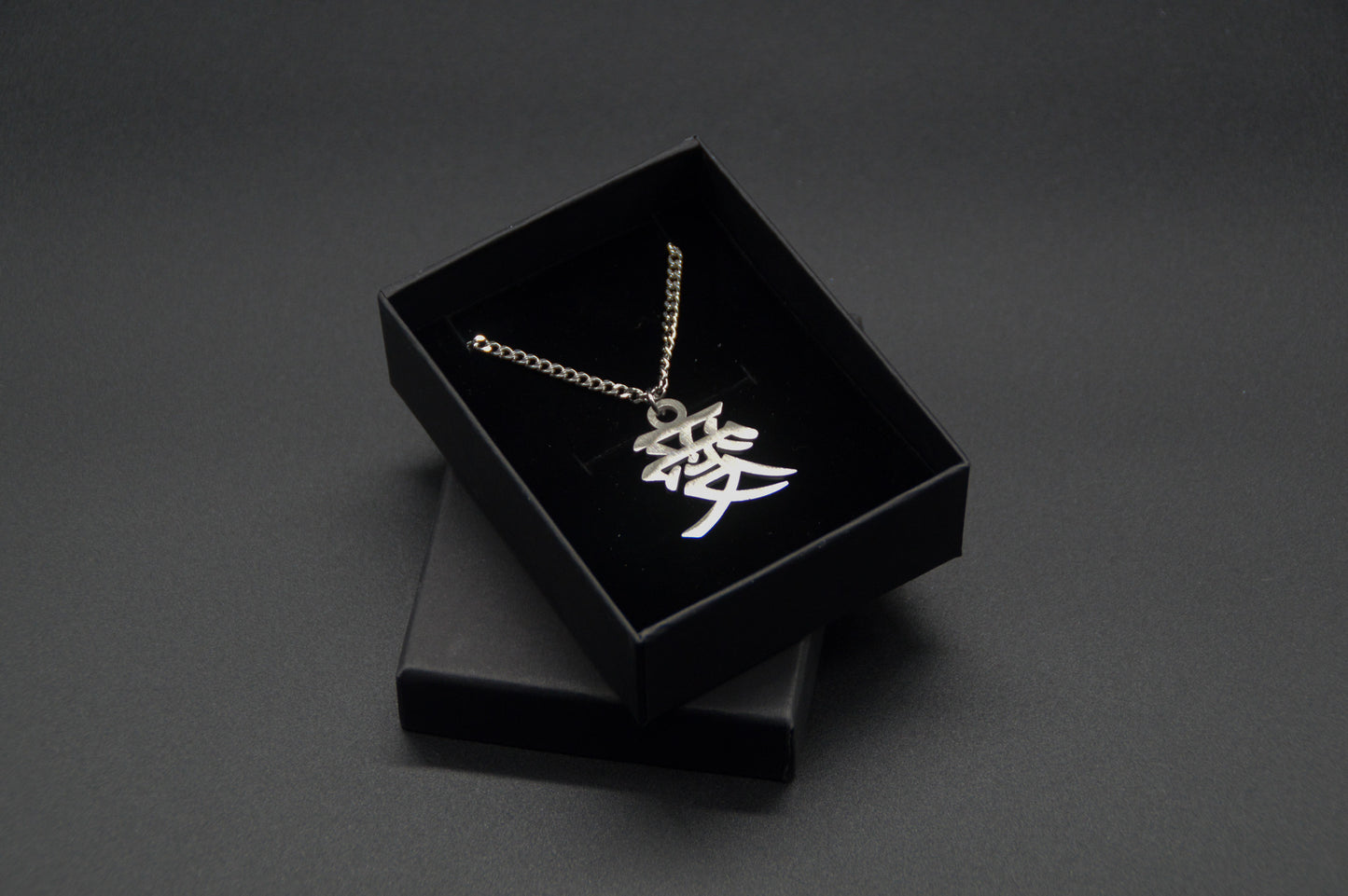 Das Japanische Schriftzeichen (Kanji für Liebe). Edelstahl Halskette mit dem Schriftzeichen 愛 als Anhänger auf einem Schwarzen Hintergrund in einer KanjiKraft Geschenkbox verpackt.
