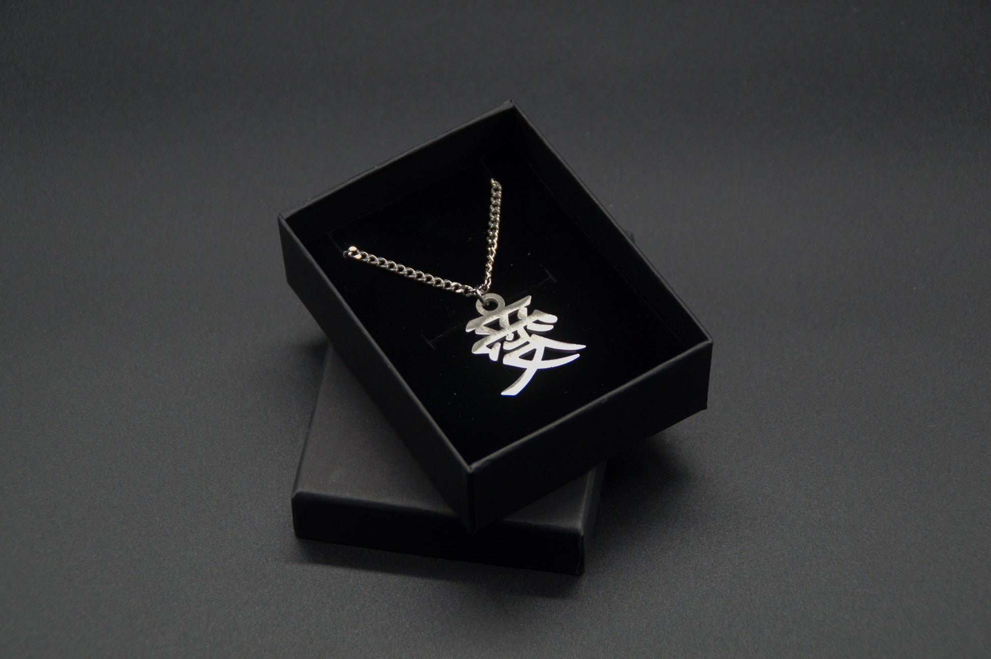 Das Japanische Schriftzeichen (Kanji für Liebe). Edelstahl Halskette mit dem Schriftzeichen 愛 als Anhänger auf einem Schwarzen Hintergrund in einer KanjiKraft Geschenkbox verpackt.