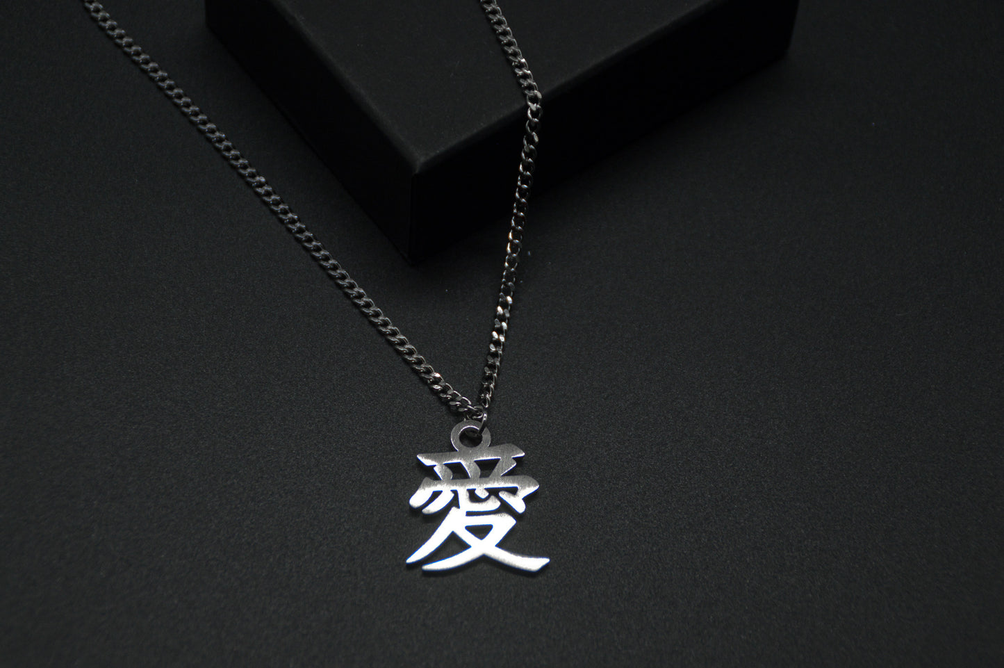Das Japanische Schriftzeichen (Kanji für Liebe). Edelstahl Halskette mit dem Schriftzeichen 愛 als Anhänger auf einem Schwarzen Hintergrund als Nahaufnahme. Die Kette liegt leicht auf einer Kante an.