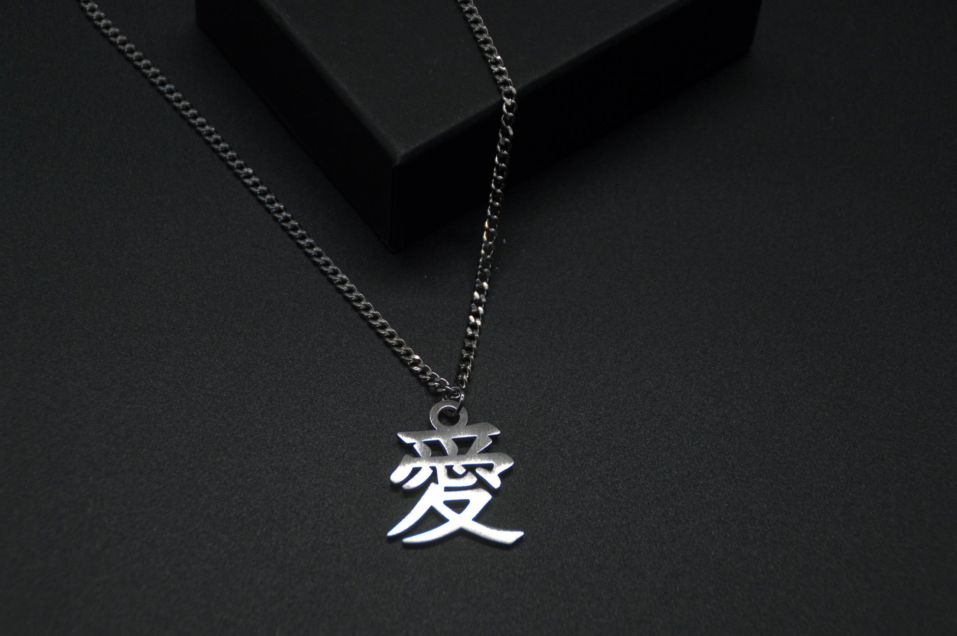 Das Japanische Schriftzeichen (Kanji für Liebe). Edelstahl Halskette mit dem Schriftzeichen 愛 als Anhänger auf einem Schwarzen Hintergrund als Nahaufnahme. Die Kette liegt leicht auf einer Kante an.