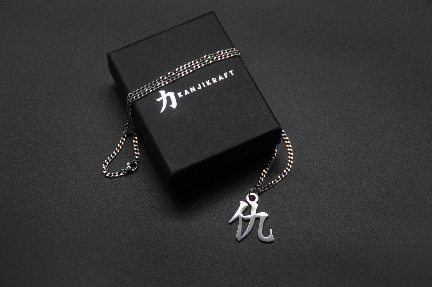 Das Japanische Schriftzeichen für Rache als Edelstahlkette auf einem Schwarzen Hintergrund auf einer KanjiKraft Box liegend