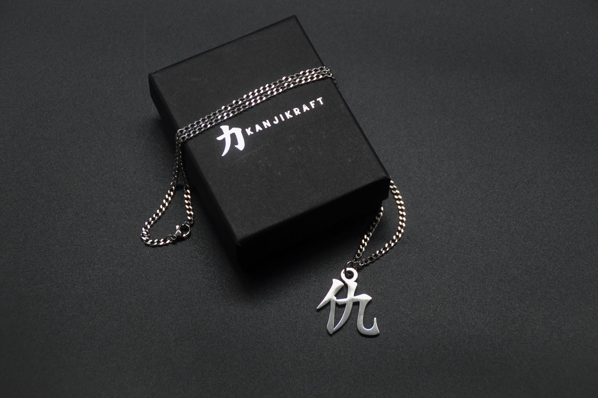 Das Japanische Schriftzeichen für Rache als Edelstahlkette auf einem Schwarzen Hintergrund auf einer KanjiKraft Box liegend
