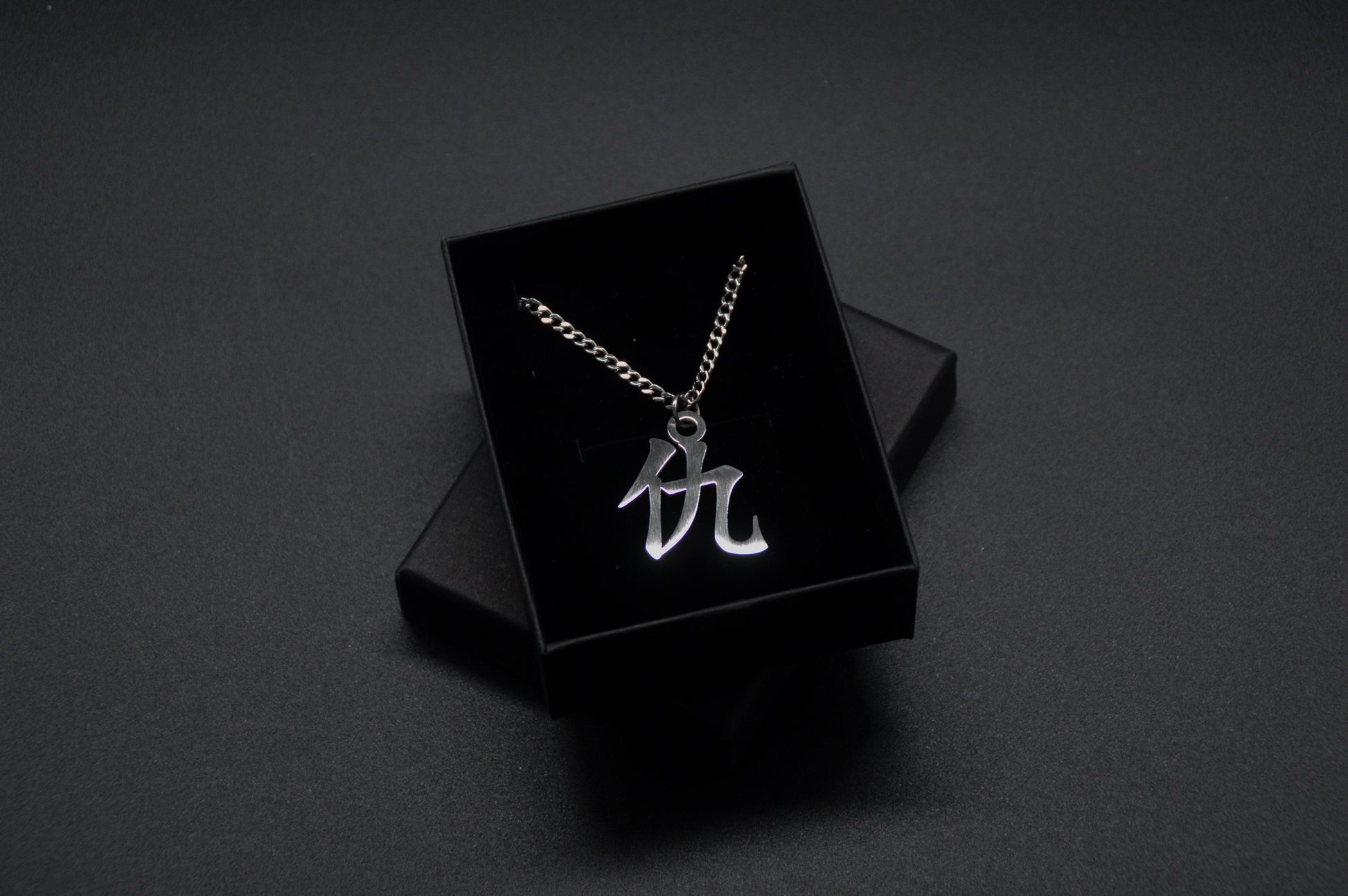 Das Japanische Schriftzeichen für Rache als Edelstahlkette auf einem Schwarzen Hintergrund in einer KanjiKraft Geschenkbox verpackt.