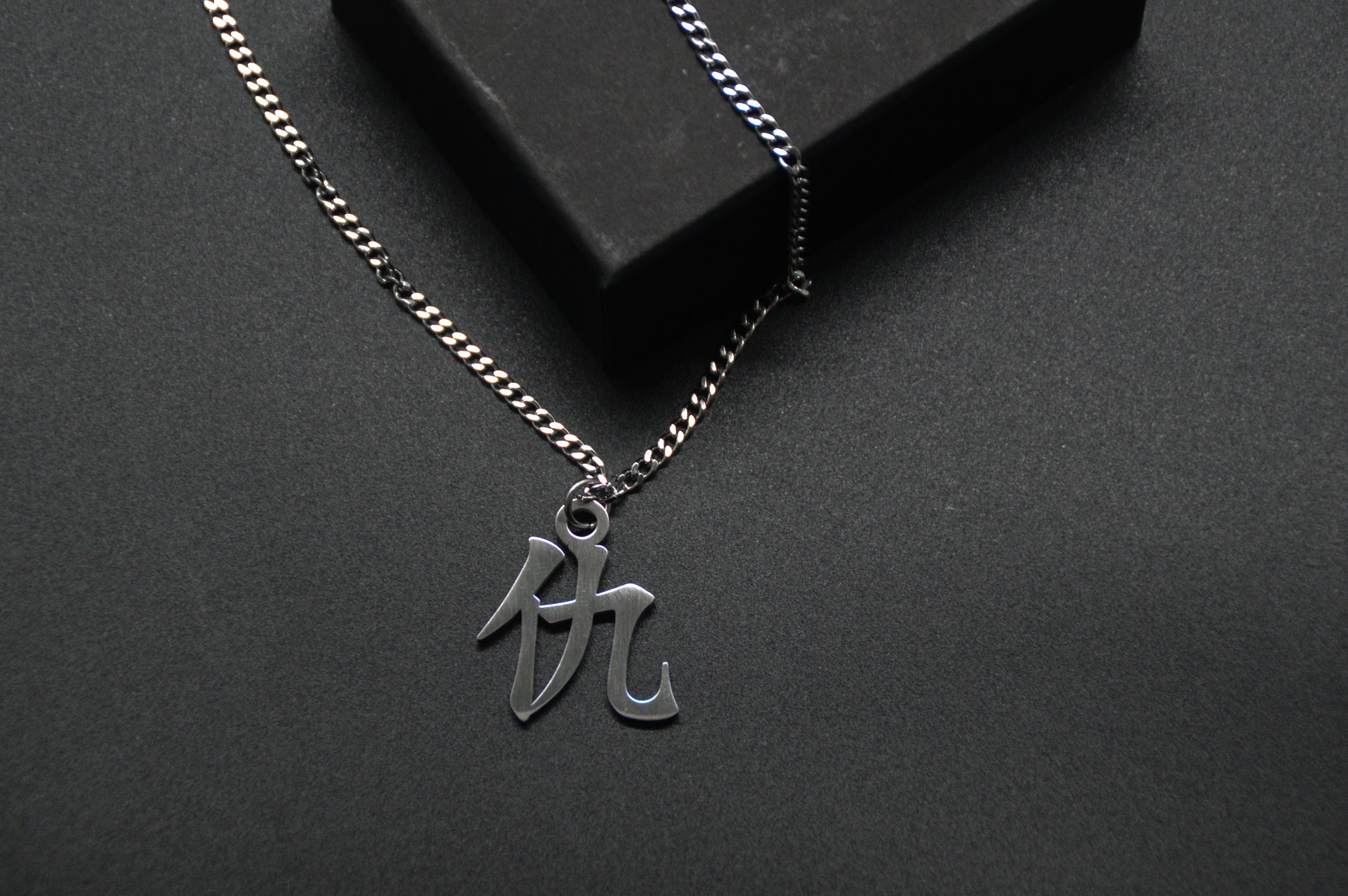 Das Japanische Schriftzeichen für Rache als Edelstahlkette auf einem Schwarzen Hintergrund. Die Kette liegt leicht an einer Kante an.